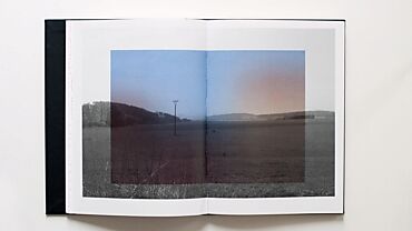 Page ouverte d'un livre d'art de Fanny Geiser. L'image montre un paysage rural avec un poteau électrique au centre, sous un ciel dégradé de bleu à orange. La moitié gauche est en noir et blanc, tandis que la moitié droite est en couleur, créant un contraste visuel.
