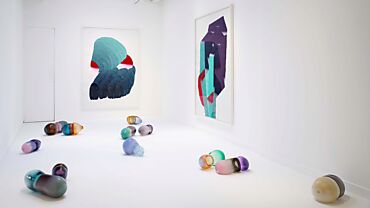 Des objets en verre colorés sont posés au sol dans une salle blanche, encadrés par deux tapisseries abstraites aux tons bleus, verts et rouges. Installation artistique d'Andrea Heller, par Parafin London.
