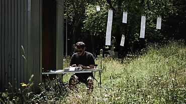 Antoine Rubin est assis dehors à une table et tape à la machine à écrire. Derrière lui, des feuilles de papier sont suspendues à une corde à linge. Photo de Pierrick Veya.