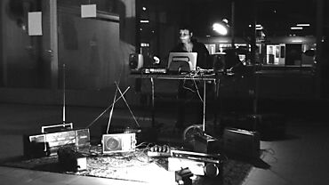 Armelle Scholl performt nachts an einem Tisch mit elektronischen Geräten und Lautsprechern. Rundherum stehen verschiedene alte Radios. Foto von Marco Rotellini.