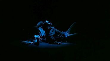 Un homme est allongé sur le sol dans une pièce sombre, éclairé par une lumière bleue. Il porte un sweat à capuche et des shorts. L'atmosphère est mystérieuse et introspective. Photo de Lucas Dubuis par Eve Chariatte.