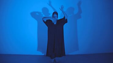 Foto von Laure Jolissaint bei einer künstlerischen Performance, in Blau beleuchtet mit deutlichen Schatten an der Wand hinter ihr, erhobene Arme und Bewegung in einem dunklen Kleid.