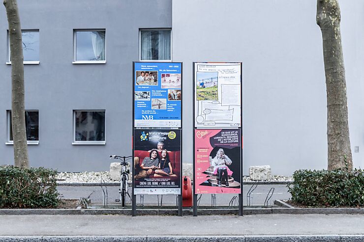 Deux panneaux d’affichage montrant quatre affiches d’institutions culturelles biennoises de différentes couleurs sont situés entre deux arbres devant deux immeubles gris.