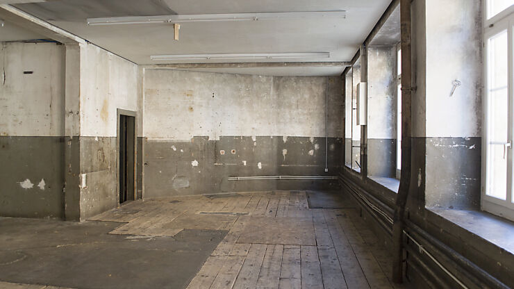 Im leeren Veranstaltungsraum sind ein alter Holzboden, unverputzte Wände und auf der rechten Seite eine durchgehende Fensterfront zu sehen.