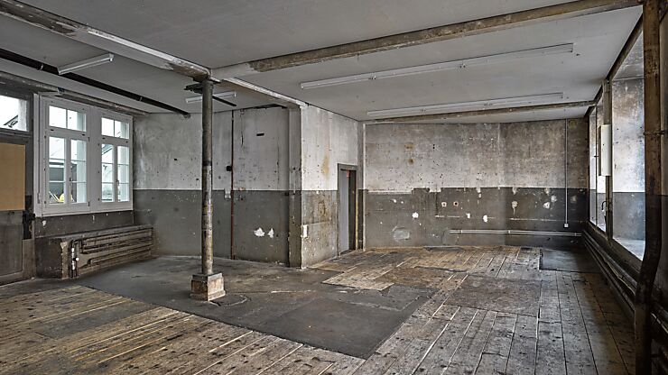 Im leeren Veranstaltungsraum sind ein Holzboden, unverputzte Wände, eine freistehende Säule und auf der linken und rechten Seite Fenster zu sehen.